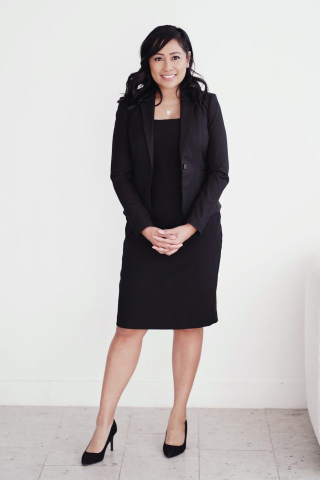 A smiling woman with dark hair stands in front of a white wall, wearing a black dress, a black blazer, and black high heels. Her hands are clasped in front of her. She is looking directly at the camera.