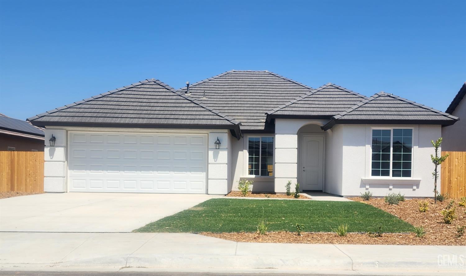 A single-story, modern-style house with a light gray stucco exterior and a dark gray tile roof. The house features a two-car garage with a white door, a small front lawn, and a paved driveway. The front entrance is recessed under an archway. A small tree is planted in the planter bed to the right of the driveway.