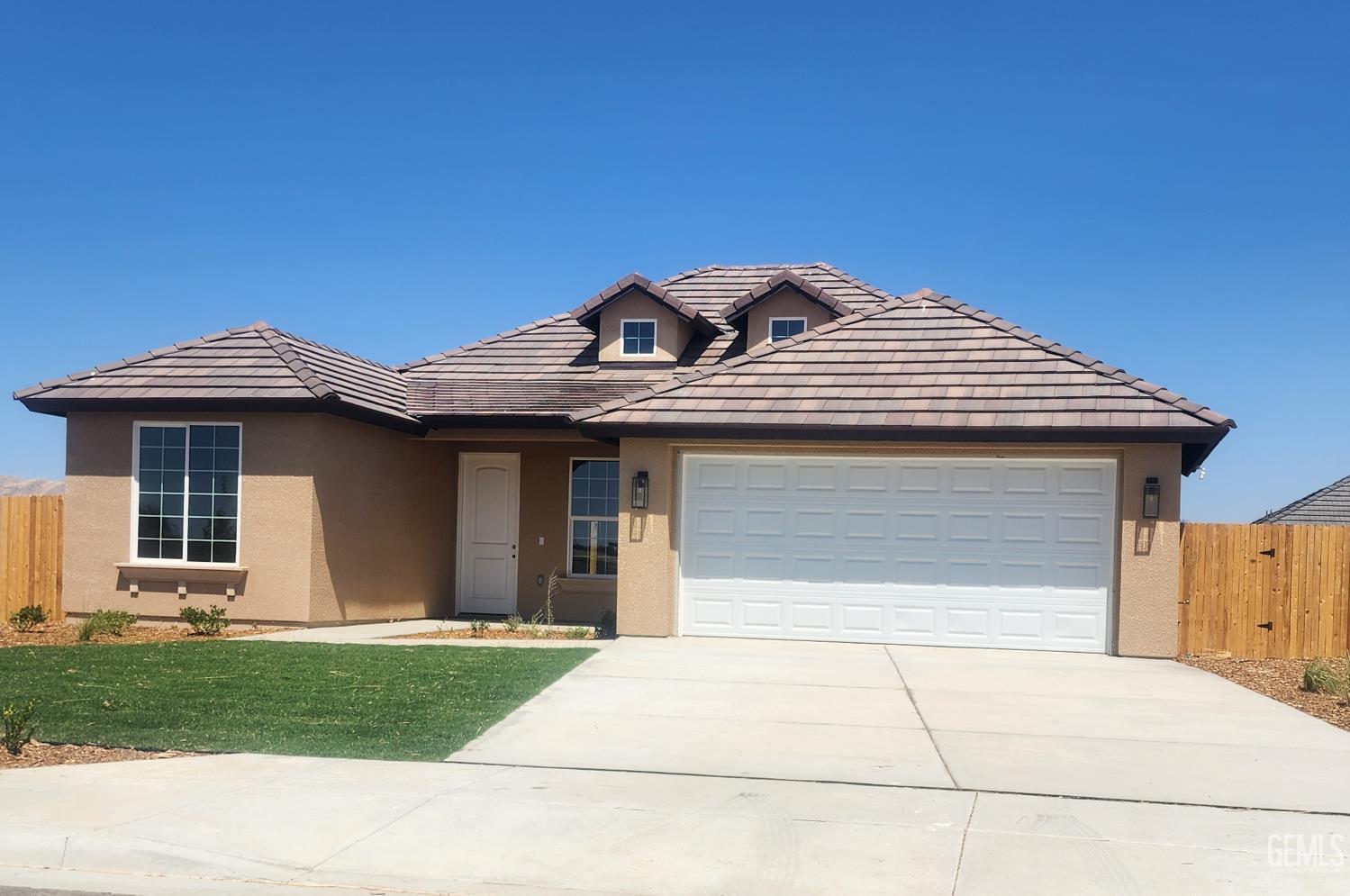 A one-story, light brown stucco house with a terracotta tile roof and a three-car garage with a white door. There are two dormer windows on the roof. A small lawn and paved driveway are visible in the foreground. A wooden fence is on the right side of the house.