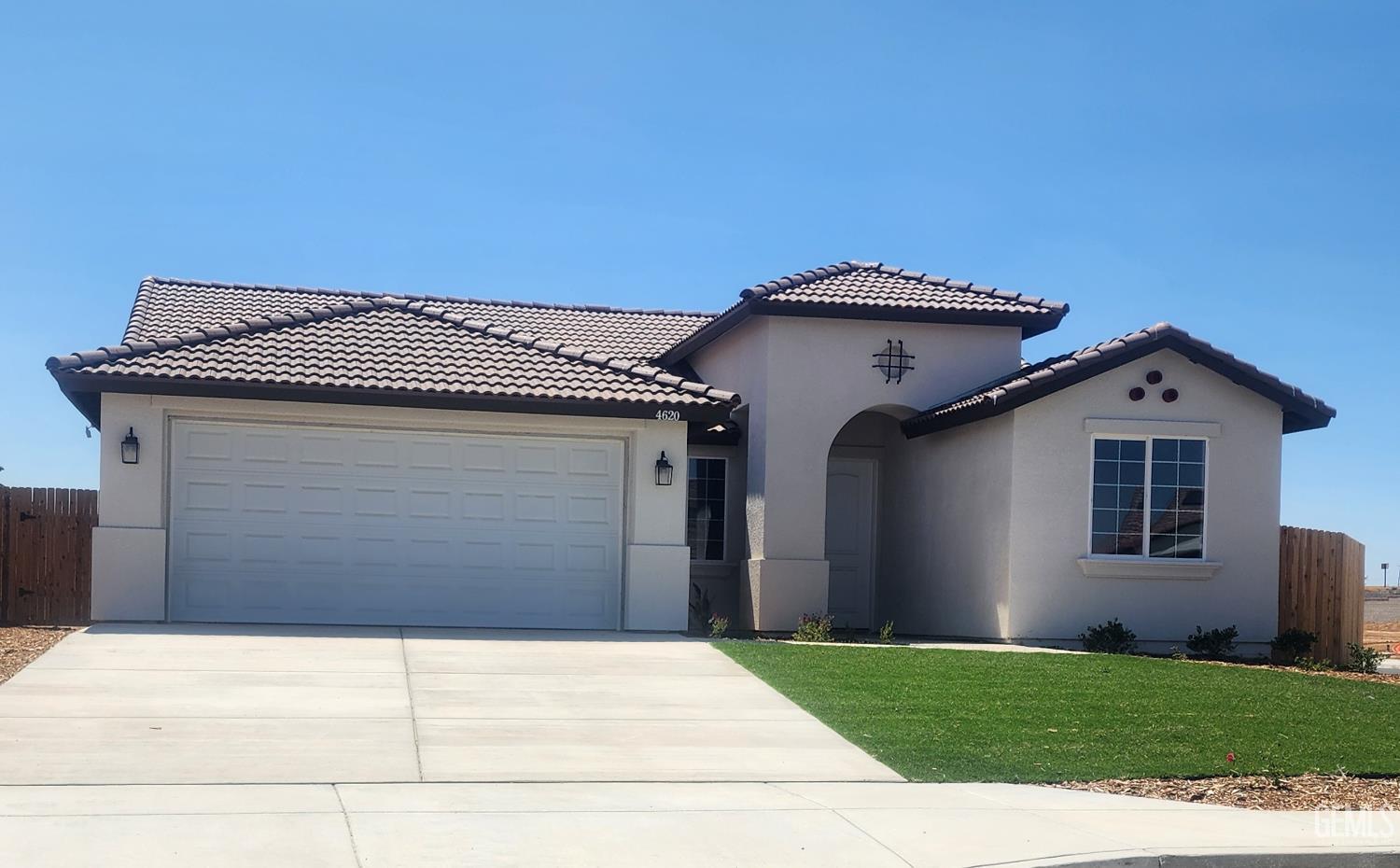A two-story, Mediterranean-style house with a light stucco exterior and a terracotta tile roof. The house has a two-car garage with a white door, a manicured lawn, and a paved driveway. The front entry is under a small archway with a decorative iron piece above it. The house number "4620" is visible above the garage door.